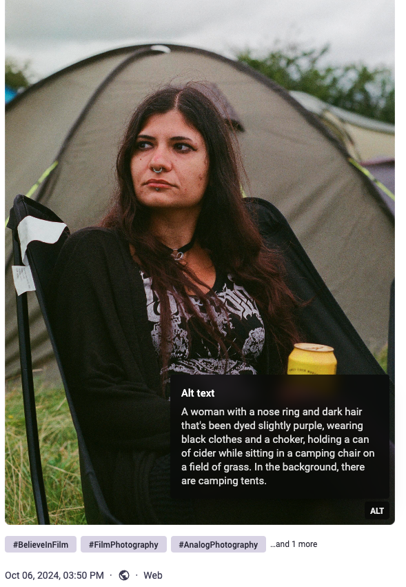 : "A woman with a nose ring and dark hair that's been dyed slightly purple, wearing black clothes and a choker, holding a can of cider while sitting in a camping chair on a field of grass. In the background, there are camping tents."