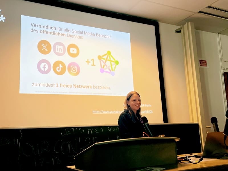 a photo of Melanie Bartos presenting at FOSDEM with a slide behind her about the concept of +1