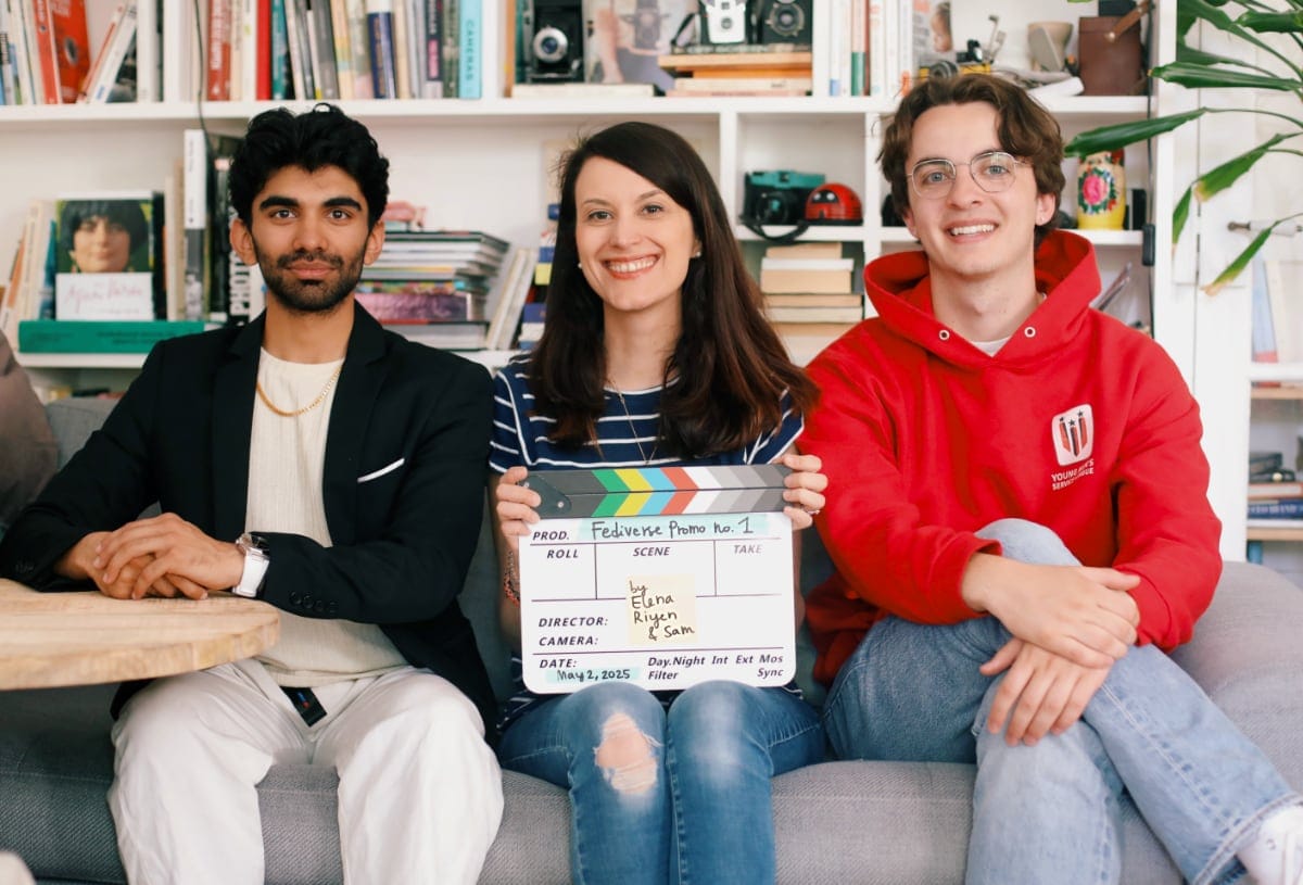 a photo of three people sitting on a couch in front of a library. A man wearing a black blazer, a woman with a striped shirt holding a clapper that reads "Fediverse promo no.1 by Elena Riyen & Sam" and a man in a red hooded sweatshirt