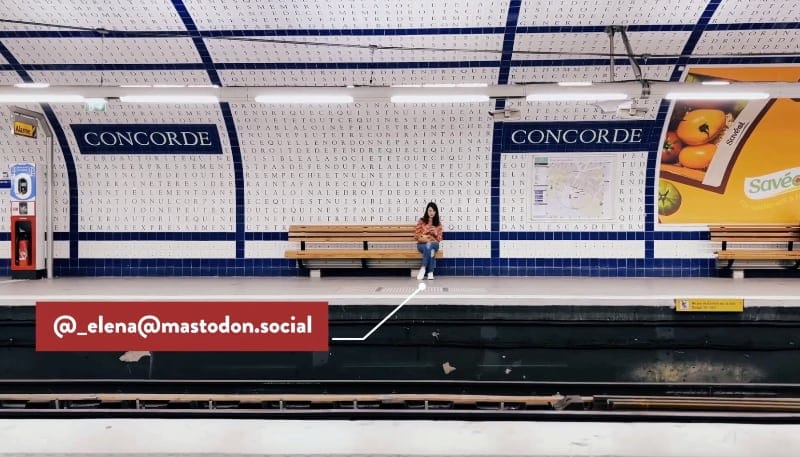 a still image from my Fediverse promo video showing me sitting on a bench inside a metro station; my Mastodon username pops up