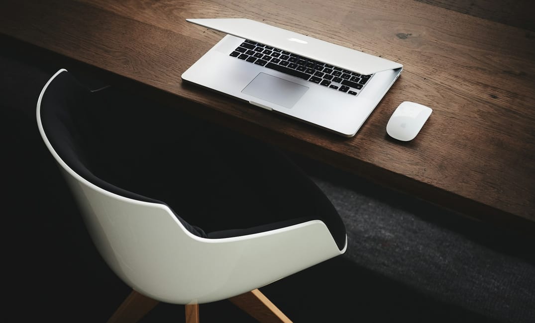 a photo showing a modern chair, a wooden desk with an Apple laptop with the lid half closed and an Apple mouse next to it